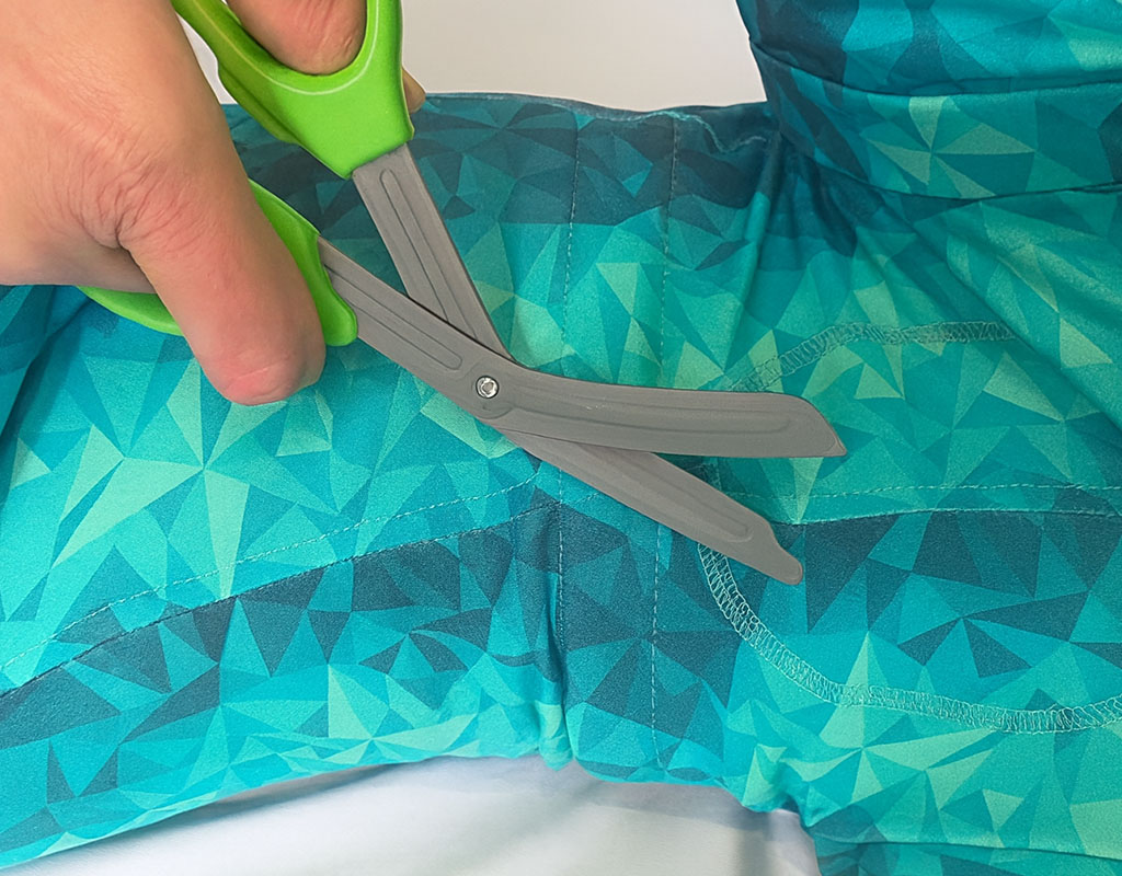 A hand holding green scissors with blunt gray blades poised to cut through a teal and blue fabric with a geometric pattern.